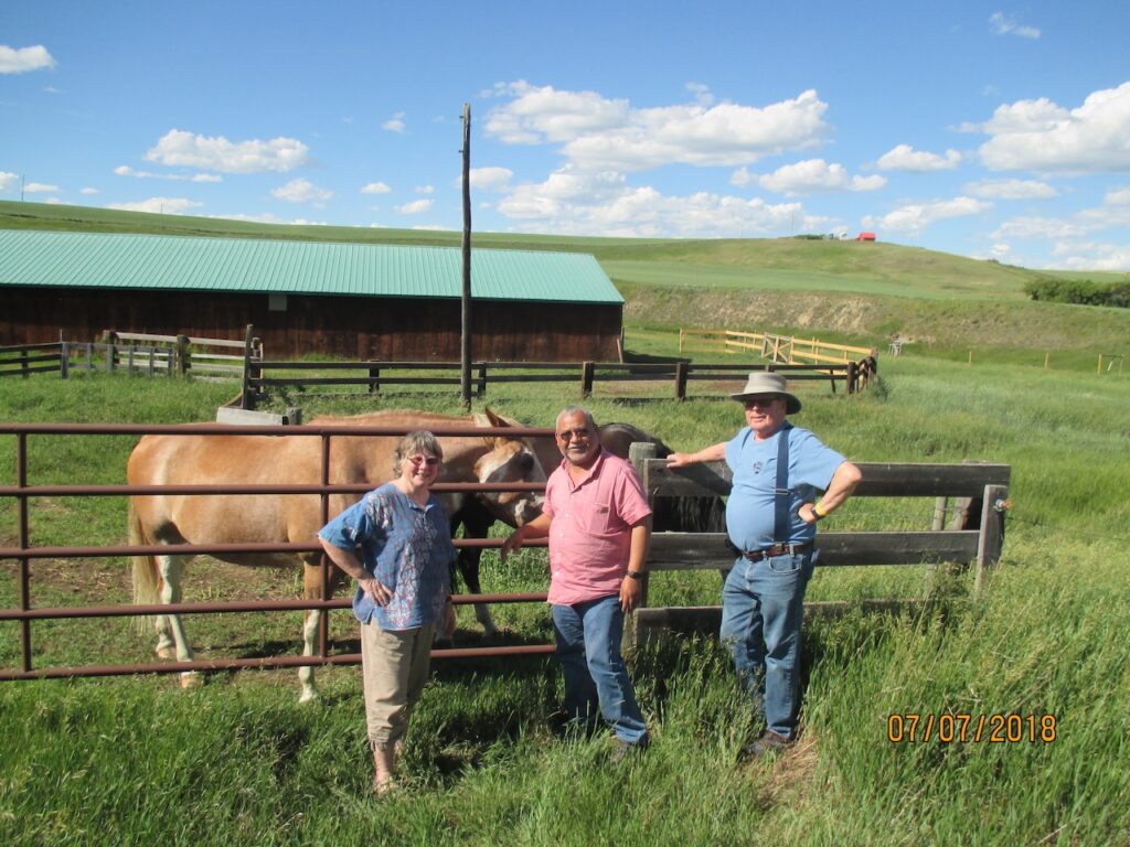 photo of two horses in corals with a long green roofed barn behind, and Melo, Anne Marie and Phil in front.