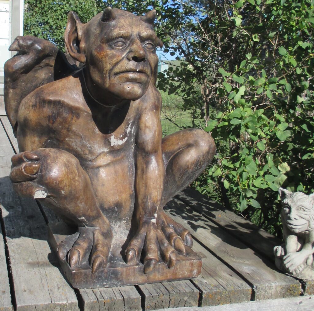 Bennedict Bray, a large dark brown concrete 200lbs gargoyle, protecting Ernst's property with the rosebud grain elevator in the background. Bennedict was dropped at the shop that sold him, causing some damages - a lost finger, a broken wing, and lots of scrapes and scratches. A smaller grey gargoyle named Lorenzo, sits to his right. The gargoyles are on a deck surrounded by arnold's red honey suckle and the Rosebud grain elevator is seen in the background.