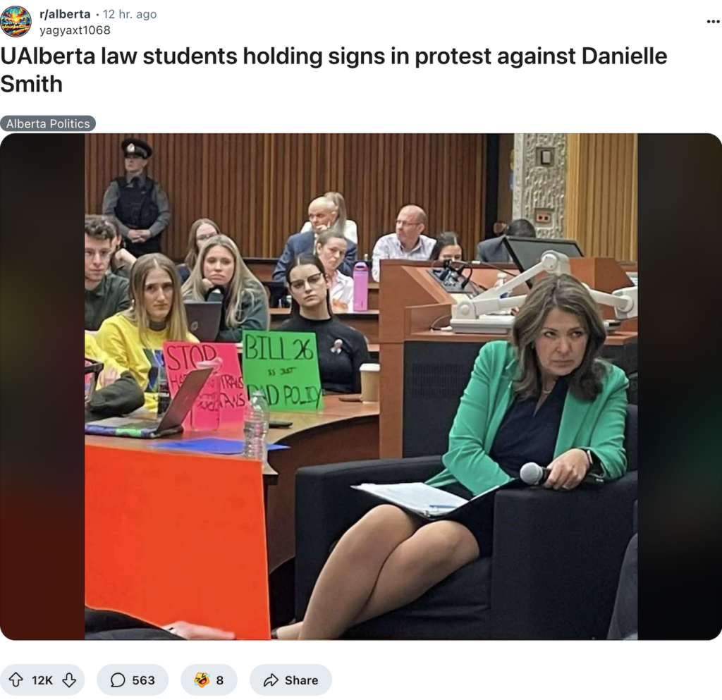 2025 03 17 photo of Alberta Premier fascist Danielle Smith unable to hide her hatred and misery, sitting in front of U of Alberta law students' protest signs against her cruel Nazi anti trans laws. Smith appears so deeply furious she looks deranged. This is a brilliant photo showing Smith's hate-filled colours