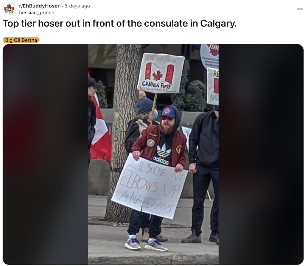 2025 03 Photo of protest against Trump's threats to destroy Canada, outside US consulate, Calgary, protester w sign, 'I'll shove my elbows up a Nazi's ass' with Canada First signs carried by protester in background