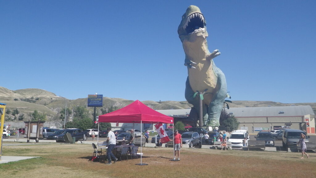 2025 08 24 Photo of the Forever Canada Petition signing station in Drumheller, Alberta (aiming to protect Canada from the minority quisling MAGAT separatists that want to destroy Canada serving Nazi Trump and handing him Alberta's riches in exchange for $500M. The station has 4 volunteers, table and chairs, a red canopy for shade, one of the volunteers wearing a red T-shirt is holding the Canadian Flag in the wind to show the Maple Leaf. A giant T-Rex dinosaur in green and yellow with wide open mouth and ragged bright white teeth and palm trees beneath it, watches over the petition station roaring, as if to tell the separatists to stand down or be eaten.