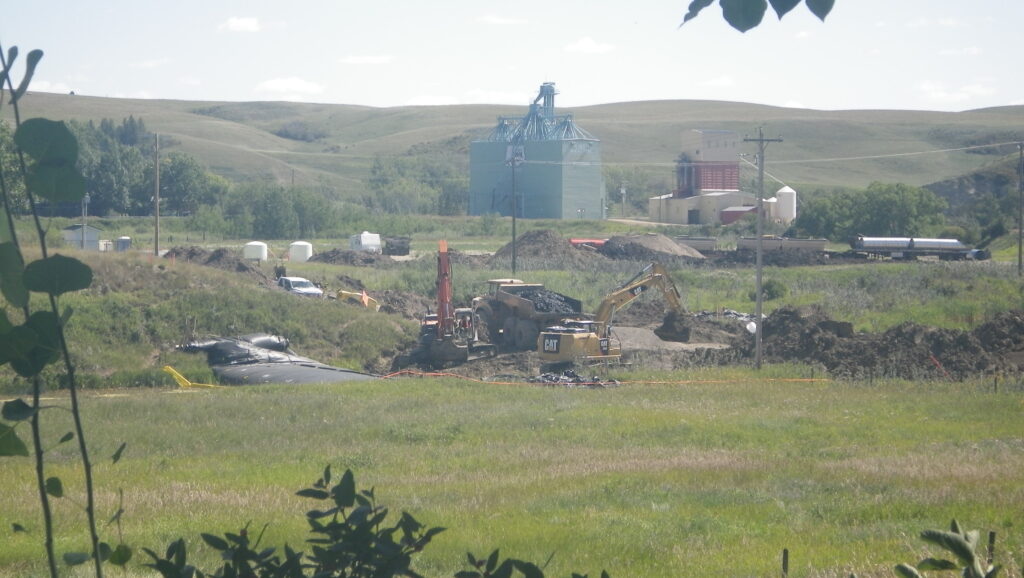 2025 08 Photo of the Old Rosebud River wetland (vital for purifying groundwater) and the road to the Ernst property being destroyed by Wheatland County. The destruction, for no valid reason (except seems to give frac'ers a water stealing parking lot) started on July 1, 2025. The fumes, dust blowing in the wind, incessant back up beepers, the stench and toxic moulds churned up from the bottom of the wetland have been going on most days 24/7. Torture. And, destroying the beauty that once was, as Albertans seem addicted to destroying. The photo was taken from Ernst's living room window. It shows a truck hauling away wetland bottom materials, piles of dirt and gravel, trucks and machinery, with the Rosebud history grain elevator and seed plant in the back ground and the coulee which amplifies the back up beepers into torture inside Ernst's home.