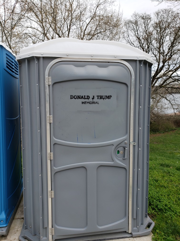 Photo of two porta-potties with trees in the background. The one on the right has a message printed on it, saying, "Donald J. Trump" Memorial. This same message is on all four porta-potties at the entrance to Banfield Park in Victoria West.