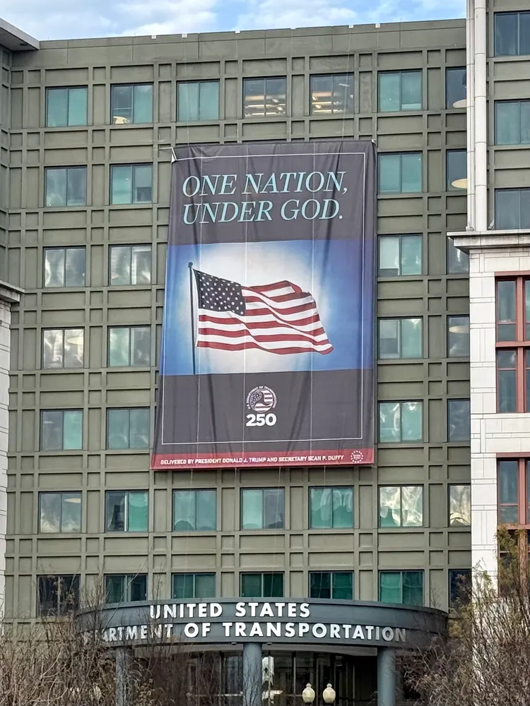 Nazi Trump Regime huge banner with american flag on US dept transportation "One Nation under God" with text bottom of banner "Delivered by President Donalt J Trump and secretary Sean P Duffy