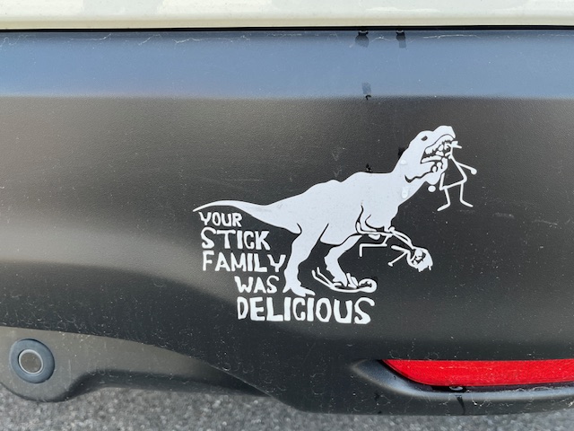 Photo of a decal showing a T Rex with a stick figure in its mouth by the head, another stick figure trampled and another in its forearms ready to nibble on. With text under its tail: "Your stick family was delicious"